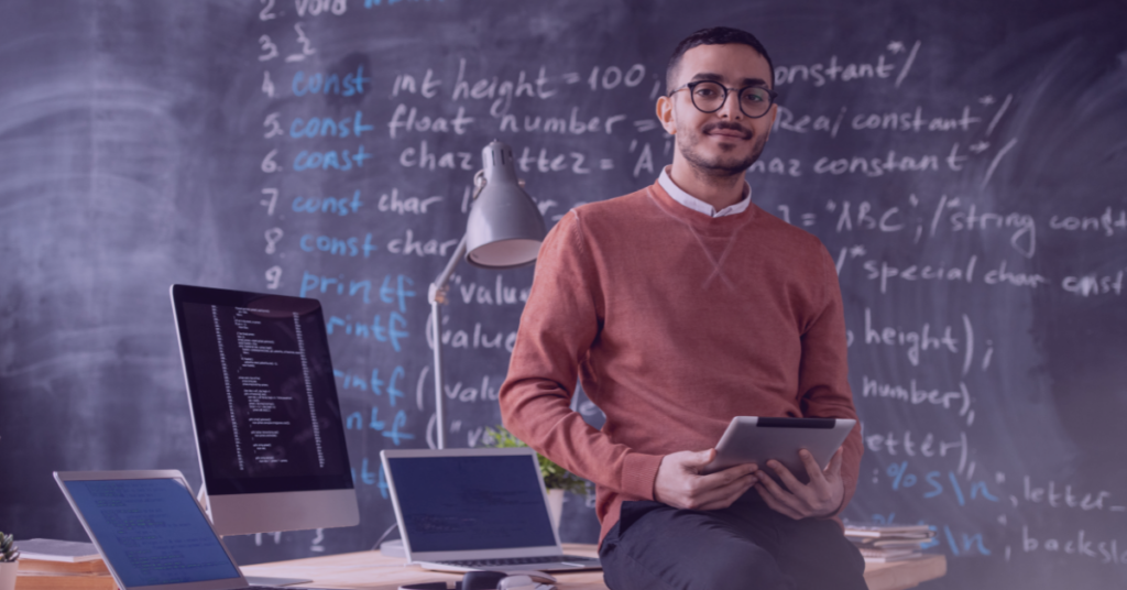 An image of a coder in front of his desk and a chalkboard full of code snippets.